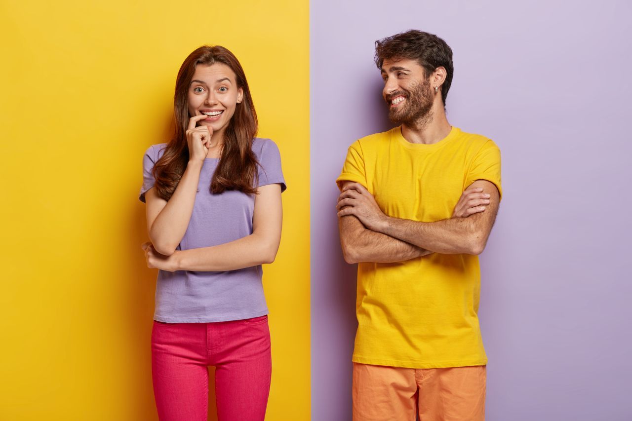 A man and a woman in an A1 German course in Frankfurt. The man looks at the woman and laughs, the woman smiles and looks slightly unsure at the camera, hand near her mouth. Background in purple, yellow, pink, and orange.