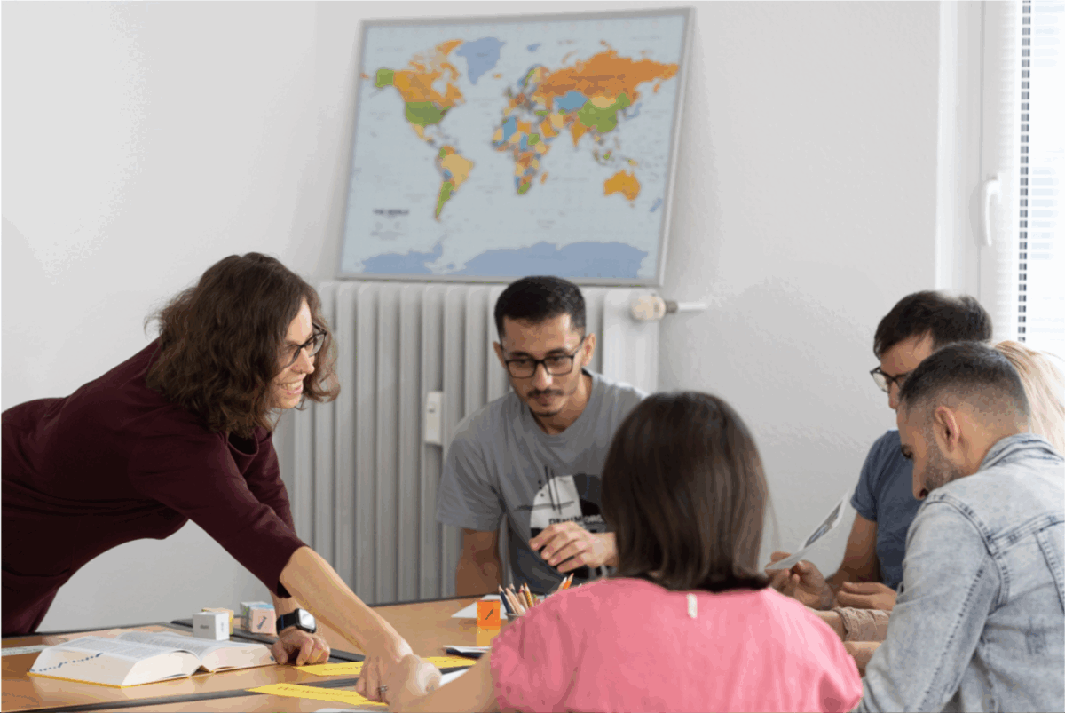 B1 Deutschkurs in Frankfurt: Fünf Kursteilnehmer arbeiten gemeinsam am Tisch, während die Lehrerin lächelnd Unterrichtsmaterial erklärt.