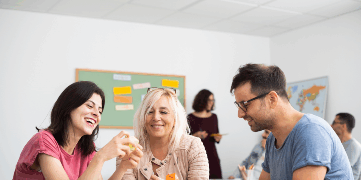 C2 Deutschkurs in Frankfurt am Main: Drei Kursteilnehmer sprechen lachend über Wortwürfel, im Hintergrund begleitet eine Lehrerin weitere Lernende im Unterricht.