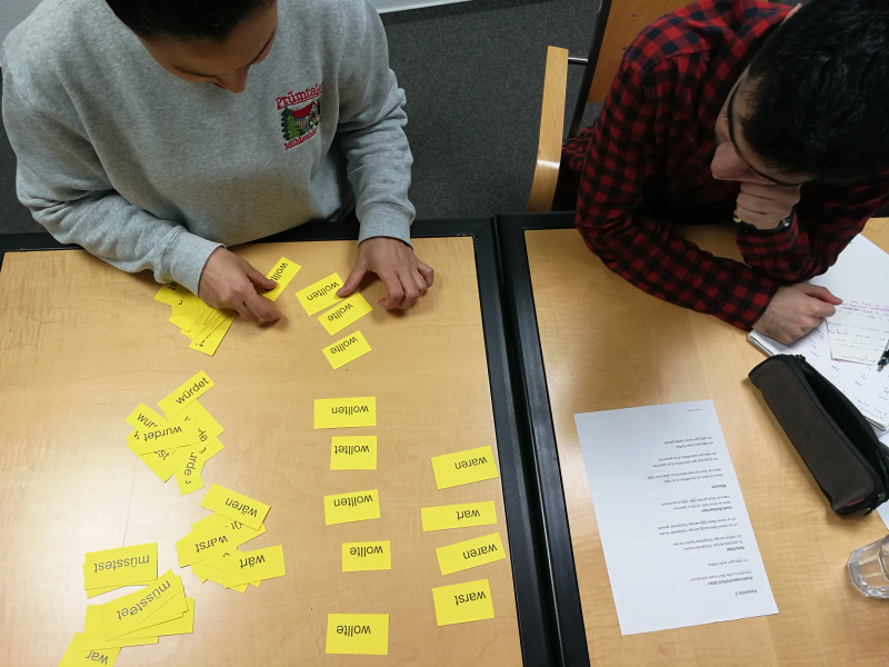 Zwei Kursteilnehmer im B2-Abendkurs in Frankfurt – eine Person legt Karten mit der Konjugation eines Verbs in die richtige Reihenfolge, um Deutsch zu üben.
