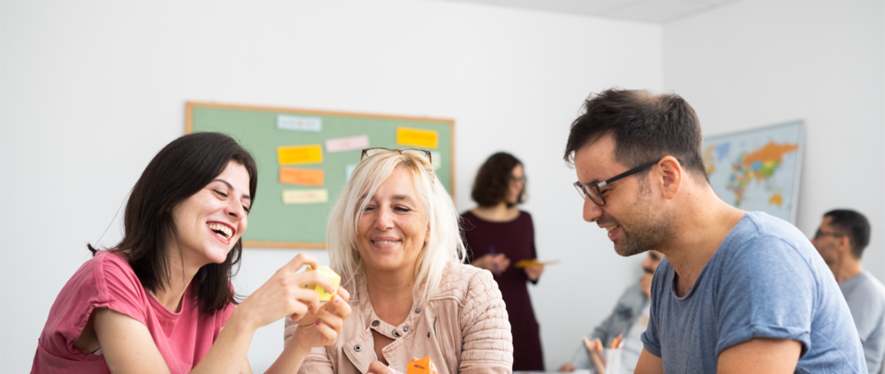 Three participants learning together in an German intensive course in Frankfurt: two smiling women and a smiling man, one woman holding a word cube. In the background, a teacher and other learners participating together in the class