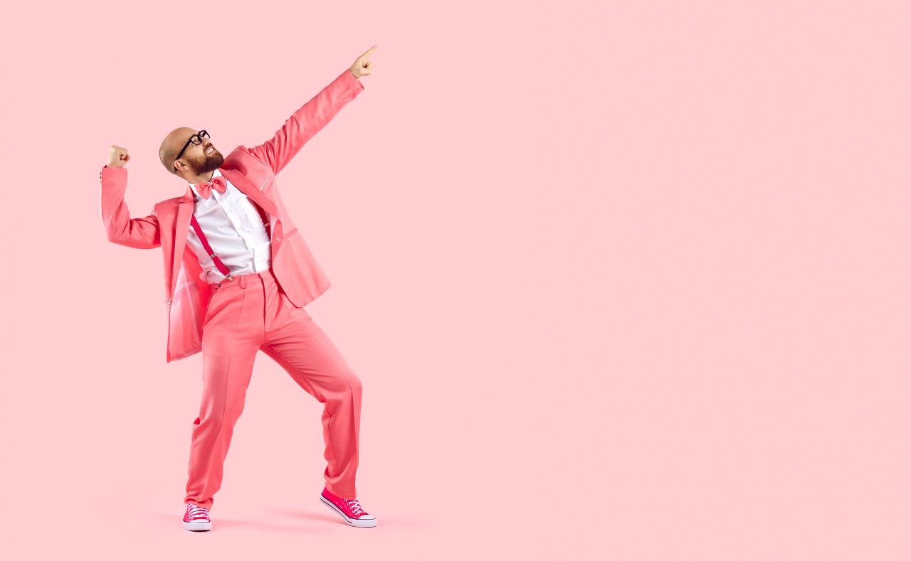 Man wearing pink suit and sneakers poses with raised arms and index fingers pointing up, as if showing “Yeah!” – symbolizing success in learning German at the language school in Frankfurt, focusing on German articles (DER, DIE, DAS).