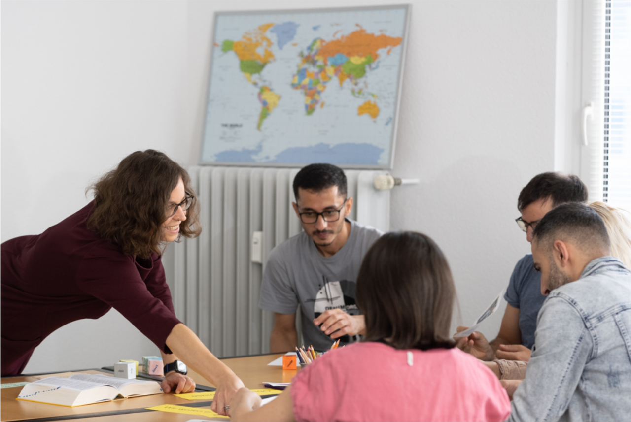 Learn German in an evening class in Frankfurt – small group with five students and a teacher leaning over the table to provide support during practice.