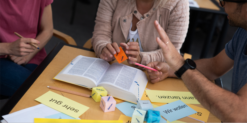 Learn German in an evening class after work in Frankfurt – close-up of hands, a connectors dice, dictionary, and sentence puzzle cards on the table, showing interactive learning.